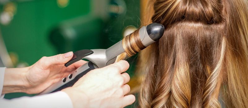 Creating curls with curling irons. Hairdresser makes a hairstyle for a young woman with long red hair in a beauty salon