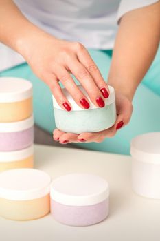 Female hands open a white body cream jar over the table with many jars of cosmetics