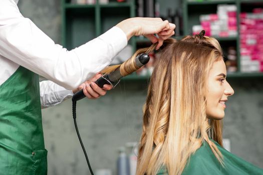 Creating curls with curling irons. Hairdresser makes a hairstyle for a young woman with long red hair in a beauty salon