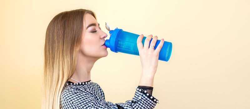 Thirsty woman with blue plastic bottle against a light background. A beautiful young caucasian businesswoman in a blouse drinking water on yellow