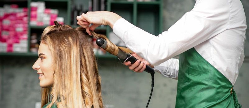 Creating curls with curling irons. Hairdresser makes a hairstyle for a young woman with long red hair in a beauty salon