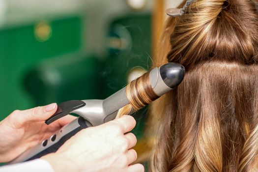 Creating curls with curling irons. Hairdresser makes a hairstyle for a young woman with long red hair in a beauty salon