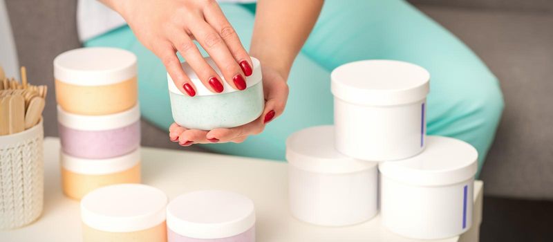 Female hands open a white body cream jar over the table with many jars of cosmetics