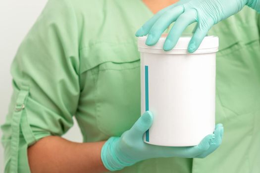 Hands in protective gloves of beautician open a white body cream jar on white background