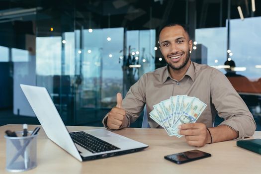 Portrait of successful young financier investor, man smiling and looking at camera, working inside modern office building, holding dollar money cash in hands and showing positive thumbs up