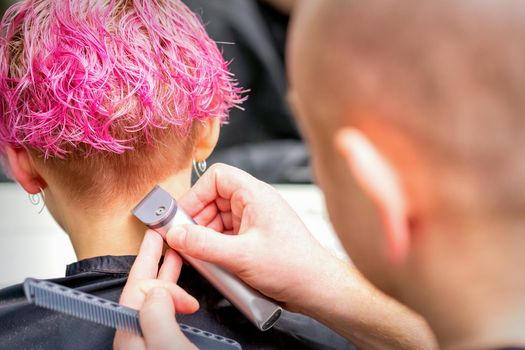 Hairdresser shaving nape and neck with electric trimmer of a young caucasian woman with short pink hair in a beauty salon