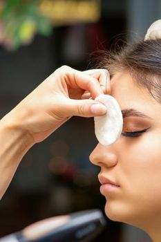 Side view of a makeup artist covers female eye with a cotton pad using airbrush making makeup foundation on her face in a beauty salon