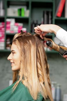 Creating curls with curling irons. Hairdresser makes a hairstyle for a young woman with long red hair in a beauty salon