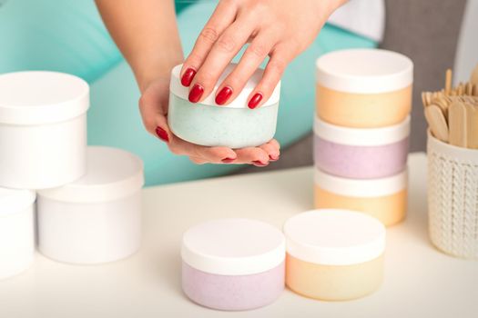 Female hands open a white body cream jar over the table with many jars of cosmetics