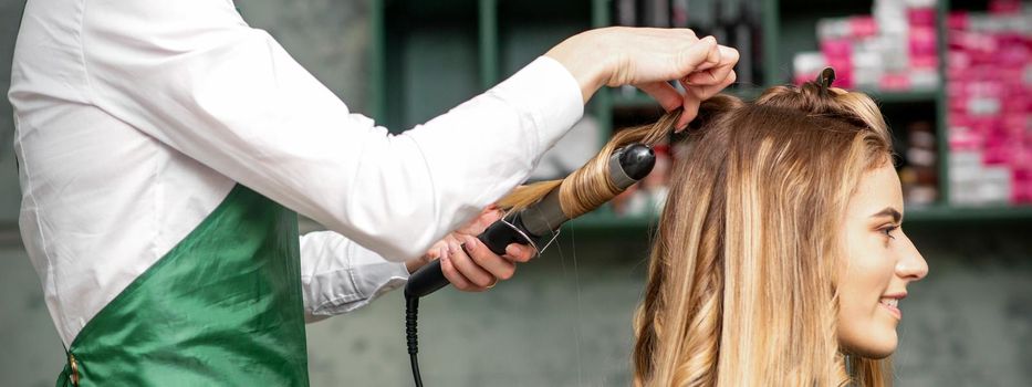 Creating curls with curling irons. Hairdresser makes a hairstyle for a young woman with long red hair in a beauty salon