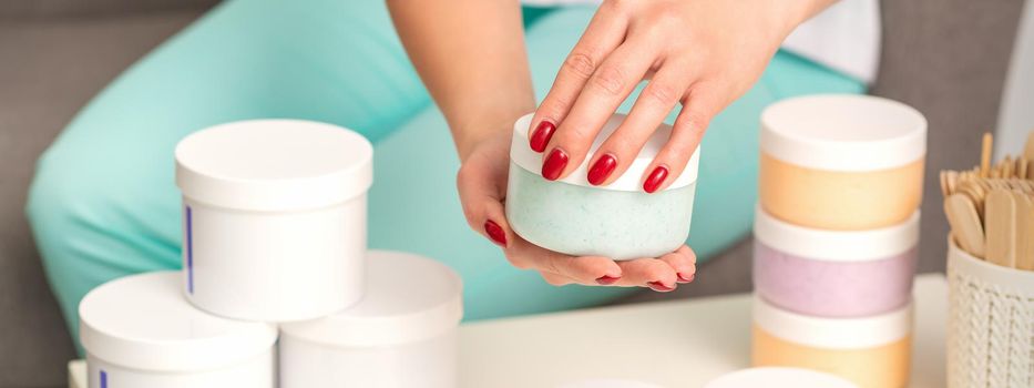 Female hands open a white body cream jar over the table with many jars of cosmetics