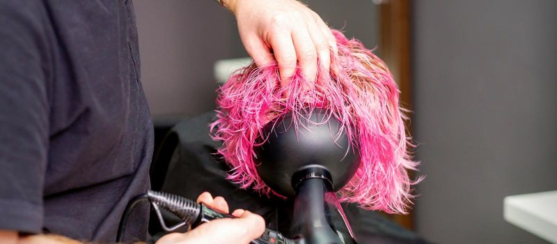 Hair Stylist making hairstyle using hair dryer blowing on wet custom pink hair at a beauty salon