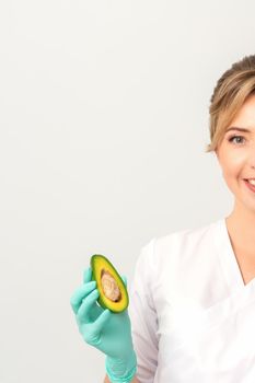 Portrait of young female nutritionist doctor with beautiful smile posing at camera holds half avocado on white background, copy space. Benefits of proper nutrition