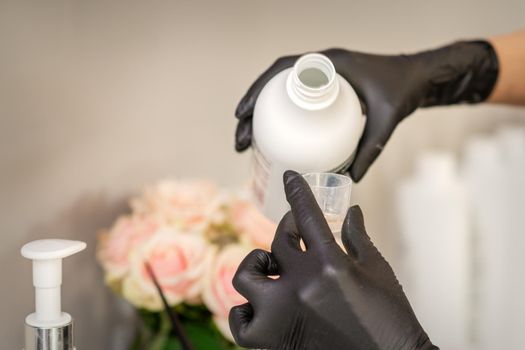 A hairdresser in black gloves is preparing hair dye with a bottle in a hair salon, close up