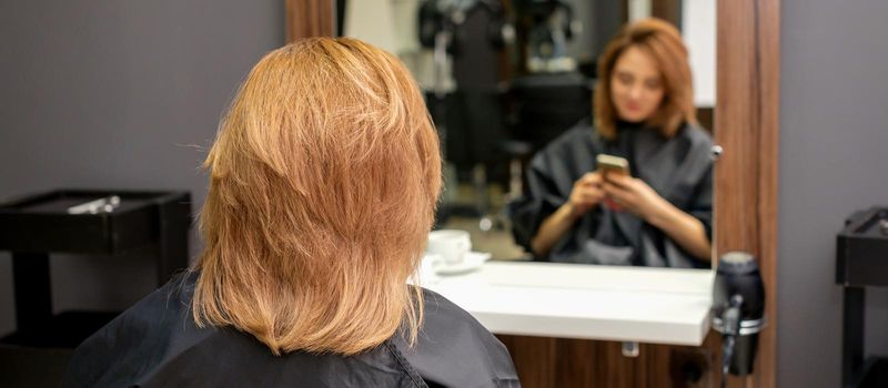 Beautiful young red hair woman using her smartphone and texting sitting in front of a mirror waiting to visit a hairdresser