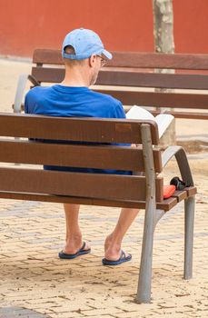 Adult man with a blue cap relaxed reading on park bench.