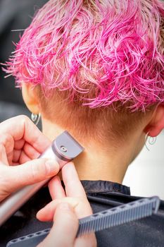 Hairdresser shaving nape and neck with electric trimmer of a young caucasian woman with short pink hair in a beauty salon