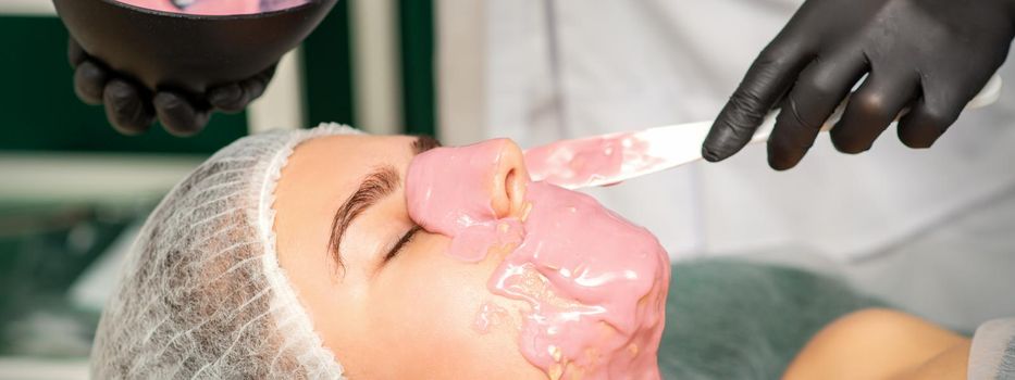 The cosmetologist applying an alginate mask to the face of a young woman in a beauty salon