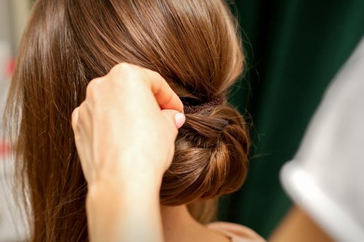 Hands of hairdresser making french twist hairstyle of an unrecognizable young brunette woman in a beauty salon, back view, close up