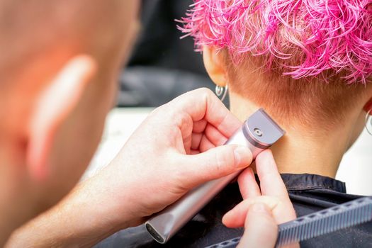 Hairdresser shaving nape and neck with electric trimmer of a young caucasian woman with short pink hair in a beauty salon