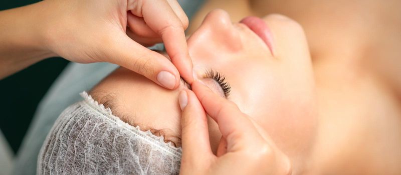 Manual sculpting face massage in the spa. Fingers of beautician make facial massage eyebrow of a young woman in cosmetology clinic, close up