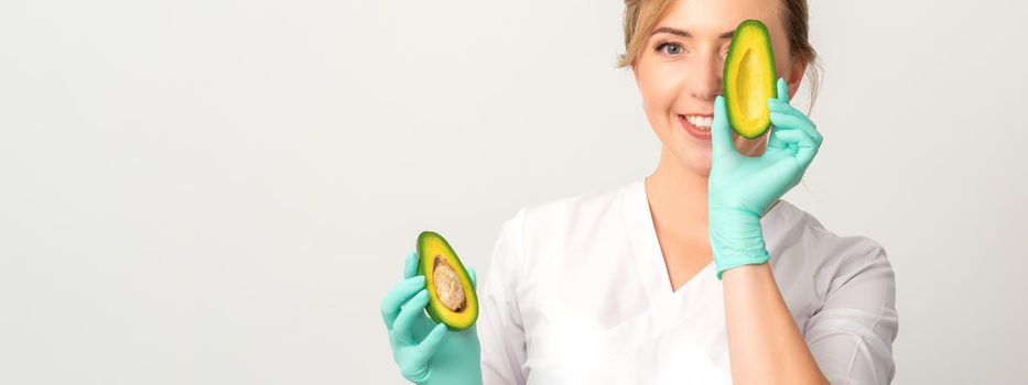 Portrait of young female nutritionist doctor with beautiful smile posing at camera hiding eye behind half avocado on white background, copy space. Benefits of proper nutrition