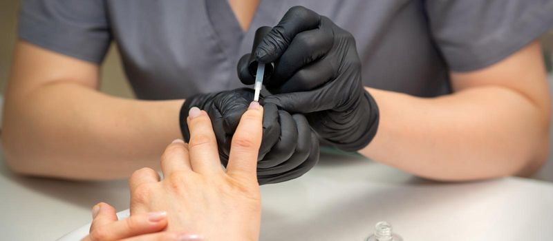 Painting female nails. Hands of manicurist in black gloves is applying transparent nail polish on female nails in a manicure salon