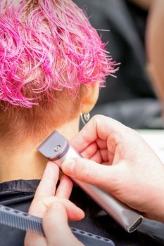 Hairdresser shaving nape and neck with electric trimmer of a young caucasian woman with short pink hair in a beauty salon