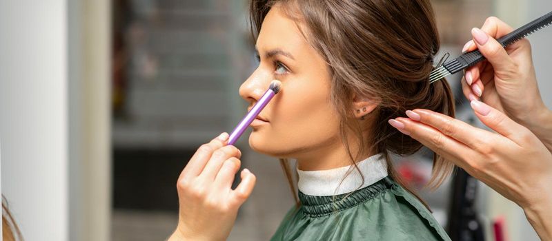 Makeup and hairstyle process. Makeup artist and hairdresser preparing young caucasian woman, working in tandem