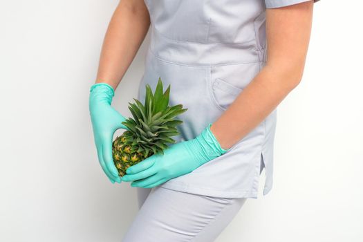 Young beautician wearing blue gloves in uniform with pineapple covers an intimate area on a white background, bikini zone depilation concept