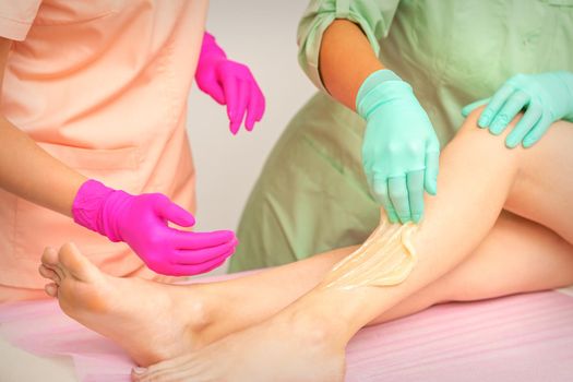 Sugaring legs. Woman legs hair removing. Hands in pink and blue rubber gloves of two beauticians apply sugar paste on female feet