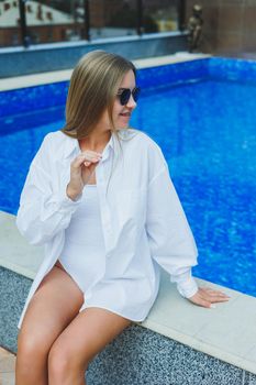 A cheerful young woman in a white swimsuit and a white shirt is sitting by the pool. A woman in sunglasses and a swimsuit. Summer vacation by the pool. Tanned girl