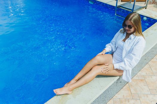 A cheerful young woman in a white swimsuit and a white shirt is sitting by the pool. A woman in sunglasses and a swimsuit. Summer vacation by the pool. Tanned girl