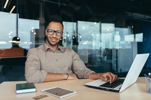 Portrait of successful businessman, man with laptop working inside modern office building, startup entrepreneur working with laptop, wearing glasses smiling and looking at camera.