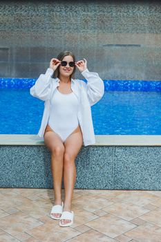 A cheerful young woman in a white swimsuit and a white shirt is sitting by the pool. A woman in sunglasses and a swimsuit. Summer vacation by the pool. Tanned girl