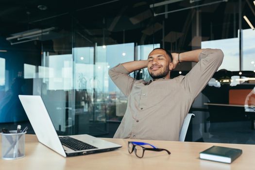 mixed race man relaxing in office with hands behind head looking out window and smiling, businessman daydreaming inside modern office building, successful investor happy with work.