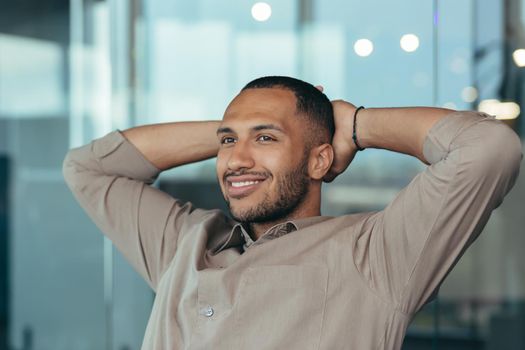 Close up photo of mixed race man relaxing in office with hands behind head looking out window and smiling, businessman daydreaming inside modern office building, successful investor happy with work