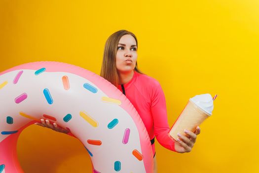A slim woman in a bright pink swimsuit on a yellow background. She is holding an inflatable pool donut. Beach fashion.