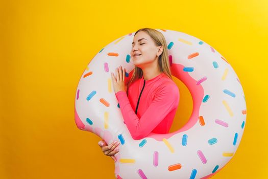 Beautiful young woman in a stylish pink swimsuit with a donut inflatable ring on a yellow background