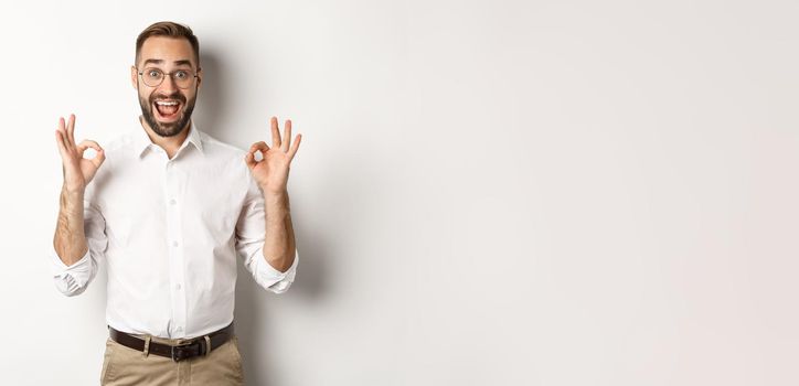 Amazed entrepreneur showing okay sign and looking happy, satisfied with product, standing over white background. Copy space