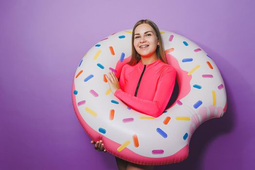 Cheerful slender woman with long hair holding an inflatable ring on a purple background. Attractive girl in a pink swimsuit