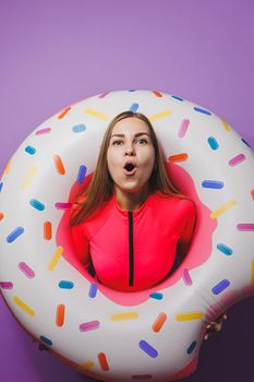 Cheerful slender woman with long hair holding an inflatable ring on a purple background. Attractive girl in a pink swimsuit