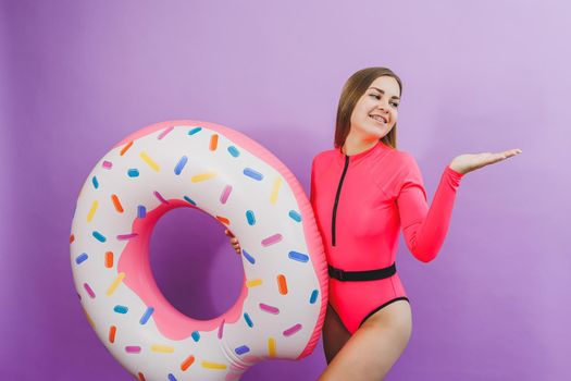 Slim young woman in a stylish pink swimsuit with a donut inflatable ring on a plain background.