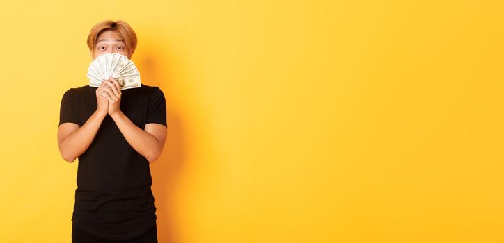 Excited lucky blond asian guy rejoicing from winning cash, holding money and looking happy, standing yellow background.