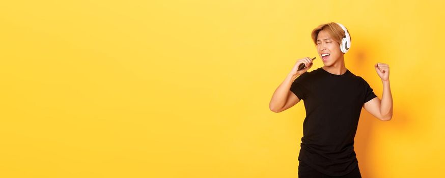 Carefree handsome asian guy in headphones, playing karaoke app, singing into mobile phone microphone, standing yellow background.
