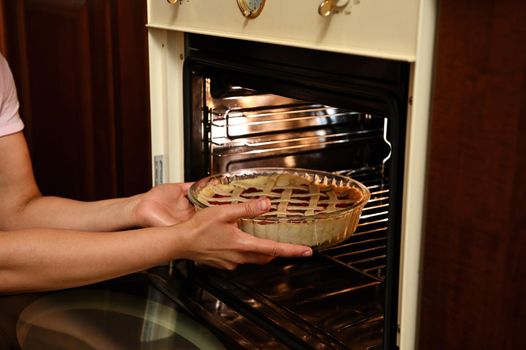 Selective focus on housewife's hands, putting homemade classic American caramelized cherry pie into oven in kitchen. Baking cherry cake.