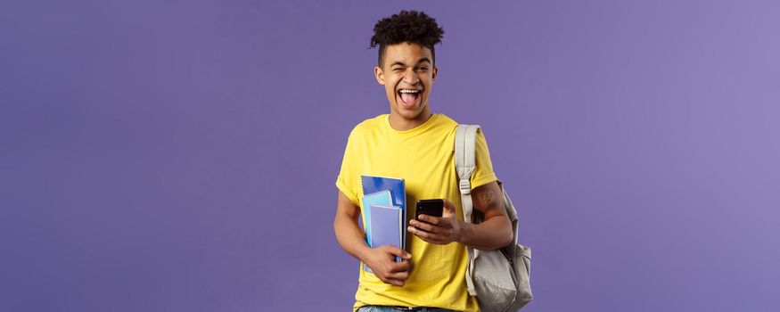 Back to school, university concept. Portrait of joyful, smiling happy man, student using mobile phone, wink at camera upbeat, holding backpack and notebooks, notes from lecture and smartphone.