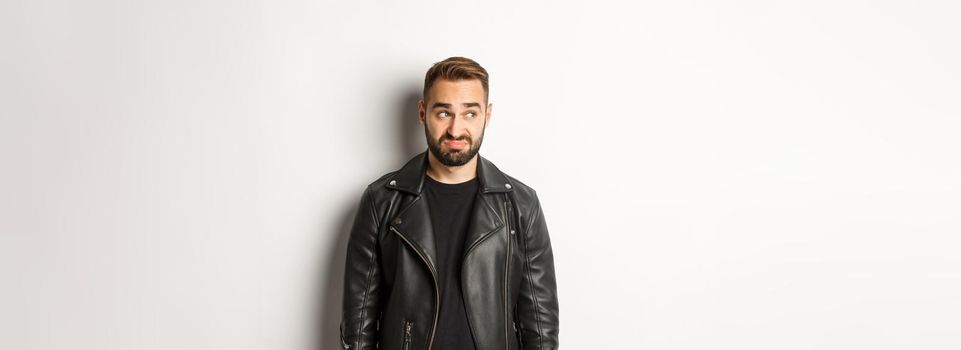 Indecisive gloomy man in black leather jacket, looking left and feeling uncomfortable, standing against white background.