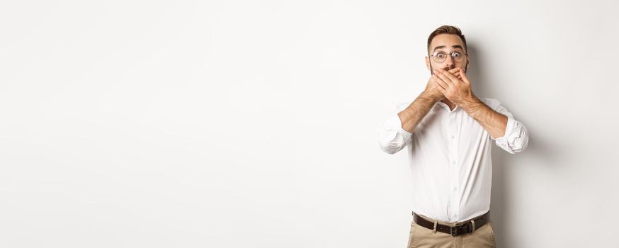 Shocked man gasping and looking at something in awe, covering mouth with hands, white background.