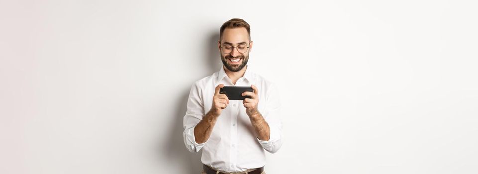 Happy businessman watching video on mobile phone, standing over white background.